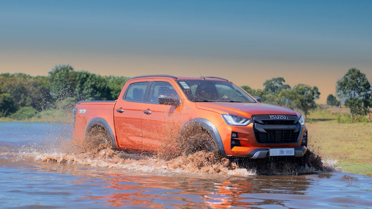 An orange Isuzu D-Max pickup truck splashing through water in a rugged terrain.